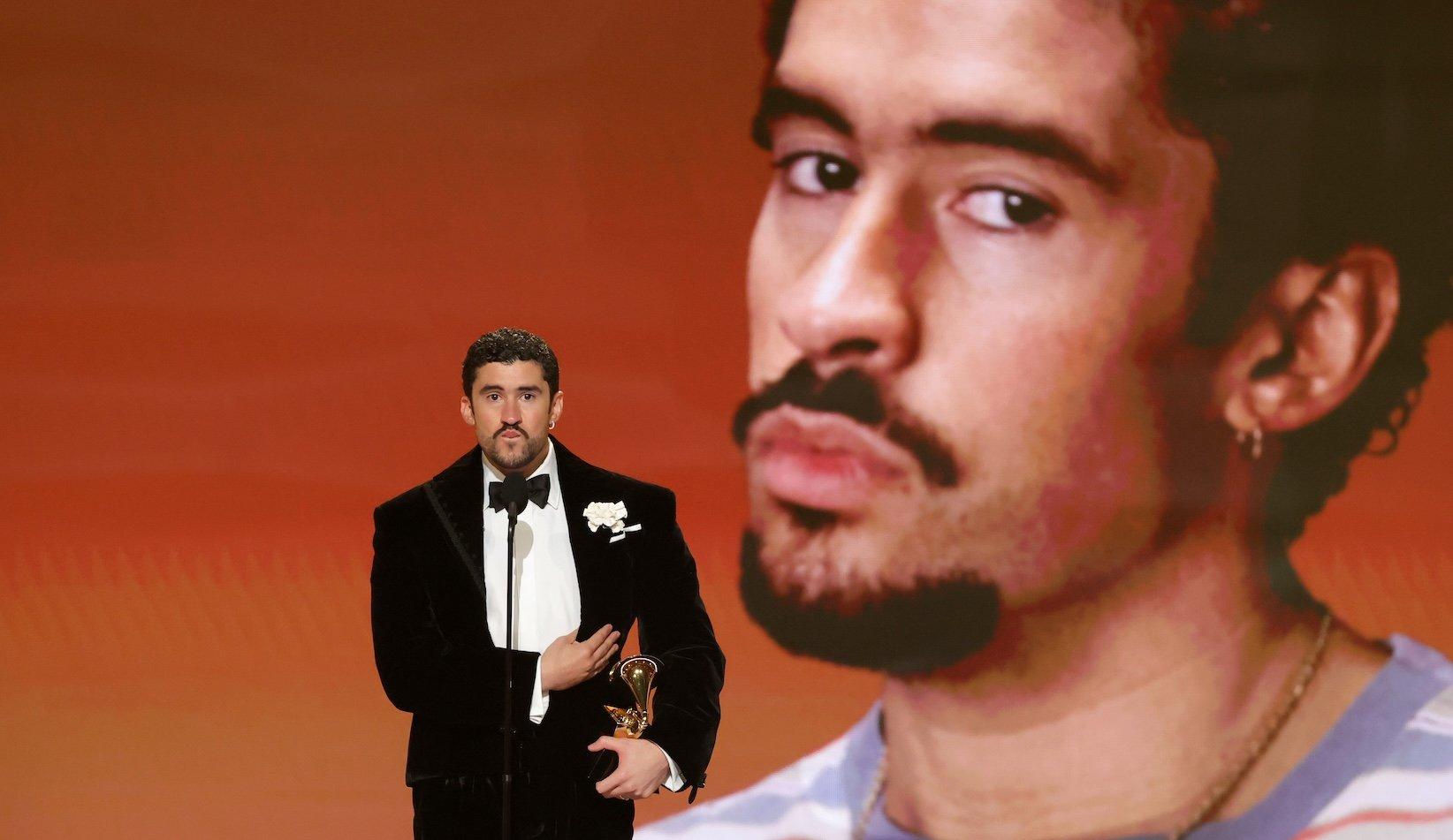 Bad Bunny wins the Grammy for Album Of The Year at the 2026 Grammys. He is standing on the Grammy stage holding his trophy and wearing a black tuxedo and white shirt.