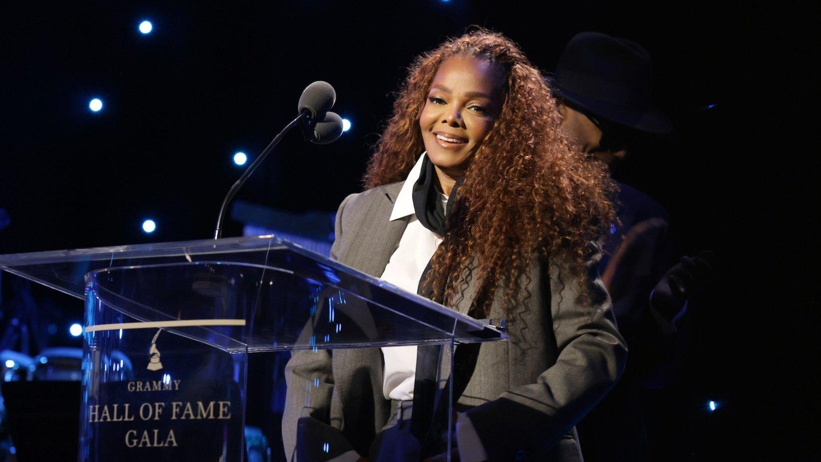 Janet Jackson accepts an honor onstage during the 2026 Grammy Hall of Fame Gala on May 8, 2026 at The Beverly Hilton in Los Angeles. Janet Jackson is wearing a grey jacket, white dress shirt, black scarf and standing in front of a microphone while smiling